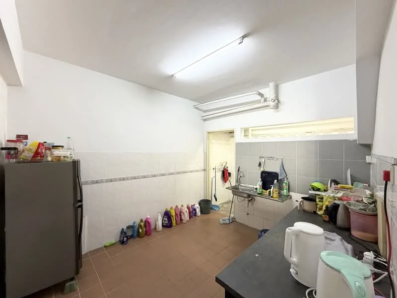 Kitchen features: white painted walls, grey tiled backsplash, black countertop, stainless-steel fridge, sink, kitchen cabinets, various cleaning supplies on floor, kettle, plastic containers, multiple bottles of cleaning products, window with grille, fluorescent ceiling lights.