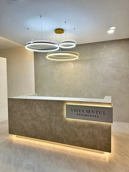 Room features: reception desk with backlighting, light-colored stone surface, modern circular pendant lights, neutral wall color, smooth wall finish, LED lighting, minimalist design, signage with 'VISTA SENTUL RESIDENCES'; style: modern.