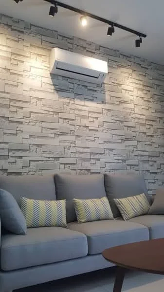 Living features: grey fabric sofa, decorative cushions with green and yellow stripes, exposed brick accent wall in light grey, recessed LED downlights, wall-mounted split AC unit, round wooden coffee table; style: modern.