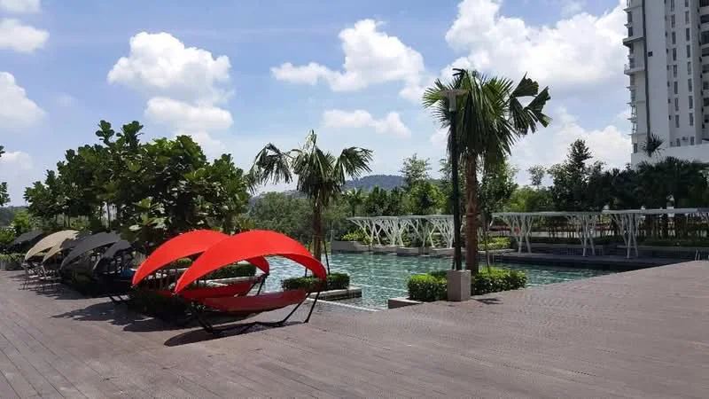 Exterior type: swimming pool deck; features: infinity pool, pool deck with loungers, landscaped garden with palm trees, manicured lawn, wooden decking, modern lounge chairs in red and beige, water feature, surrounding greenery, urban skyline in background; style: modern.