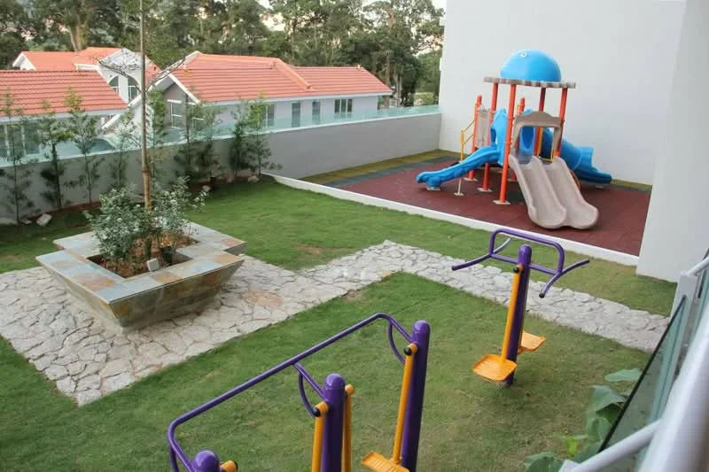 Exterior type: landscape garden; features: playground with slides and climbing structure, green lawn, stone-paved walkway, outdoor fitness equipment, decorative planter, fenced area, surrounding trees, residential buildings in background; style: modern.