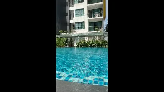 Exterior type: high-rise condominium; features: infinity pool, pool deck with loungers, landscaped garden with greenery, glass curtain wall facade, balconies visible on upper floors, tiled pool edge with blue mosaic pattern; style: modern.
