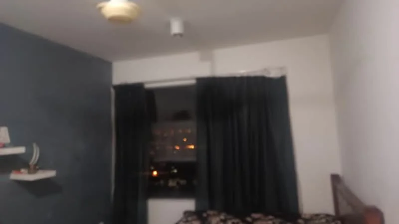 Room features: dark blue accent wall, white painted walls, floor-to-ceiling window, blackout curtains, ceiling fan, bed with patterned bedding, floating shelf, nightstand; view: city.