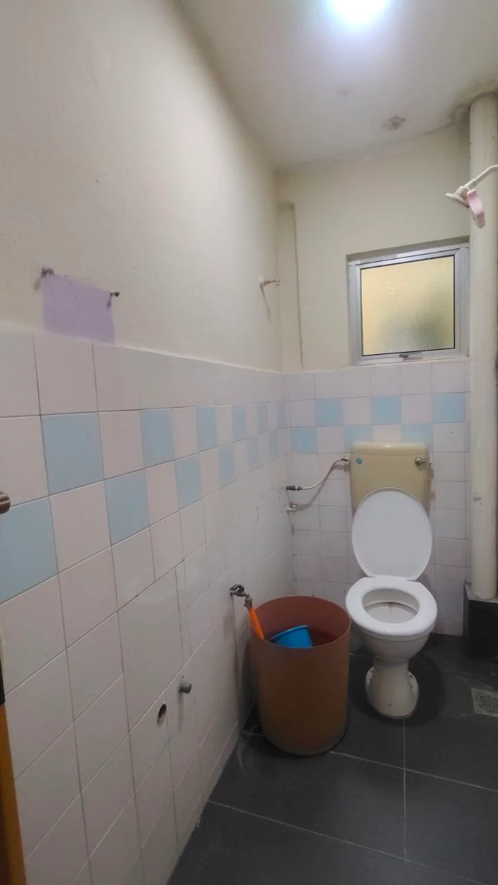 Bathroom features: white toilet, single-sink vanity, light blue and white tiled walls, grey tile flooring, window for natural light, exposed plumbing, bucket, cleaning supplies, light fixture on ceiling.
