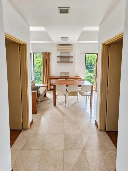 Dining features: white painted walls, beige polished tile flooring, high white ceiling, recessed ceiling light, square ceiling vent, wall-mounted split air conditioner, wooden dining table, 4 dining chairs, built-in wall shelves, small wall niche with outlet, large windows on both sides, golden-brown curtains, adjacent living area visible, wood trim around doorways, natural greenery view through windows; style: contemporary; view: garden.