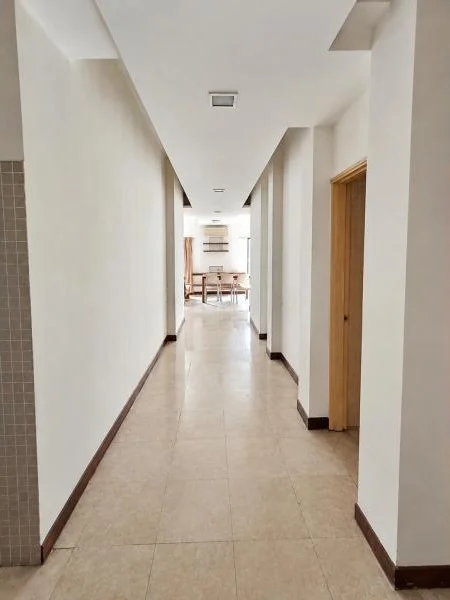 Hallway features: white painted walls, beige ceramic tile flooring, flat white ceiling, recessed ceiling lights, dark brown skirting boards, wooden door frames, multiple door openings along corridor, long narrow corridor layout, tiled accent wall at left edge, bright natural light from end of hallway; style: contemporary.