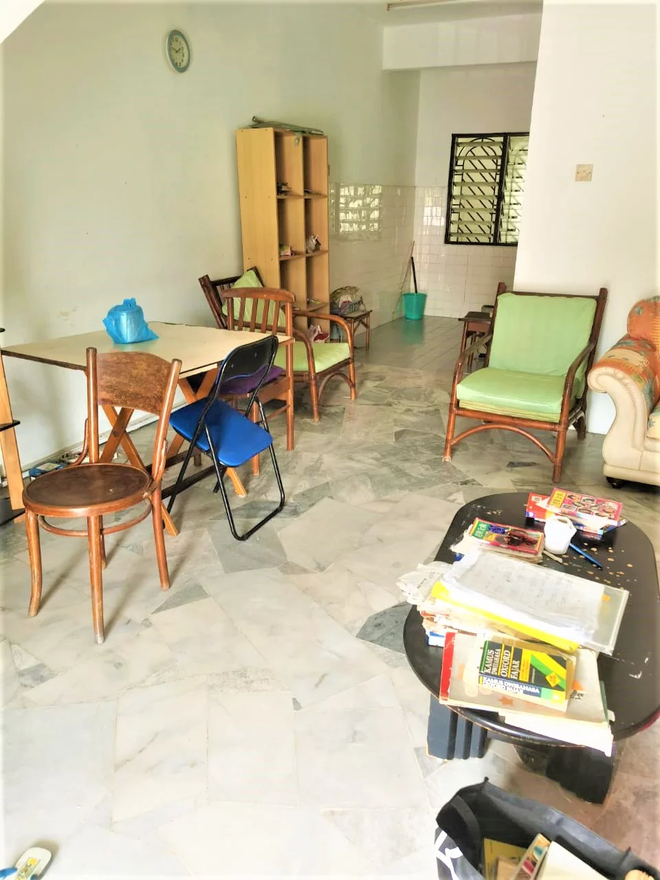 Living features: marble flooring, white painted walls, ceiling fan, wooden dining table, 4 wooden chairs, 1 folding chair, 2 green upholstered armchairs, sofa with floral upholstery, coffee table, decorative items on coffee table, wall clock, window with grille, open layout.