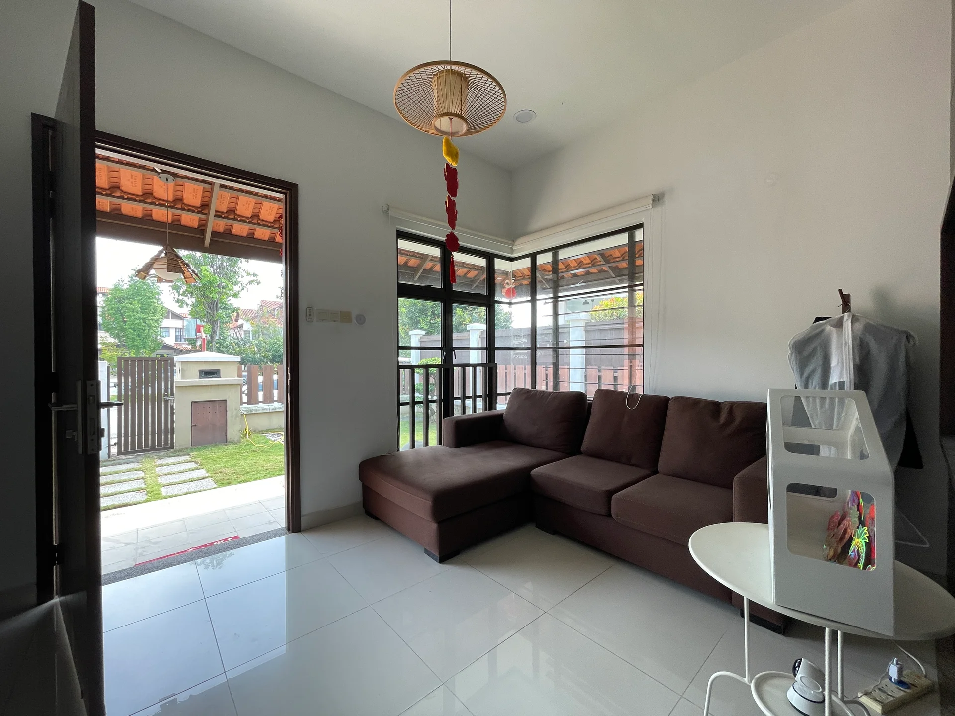 Living features: brown fabric sectional sofa, white walls, glossy white tile flooring, large window with sheer curtains, door leading to outdoor area, decorative pendant light, small round white table, coat rack with clothing, greenery visible outside; style: modern; view: garden.