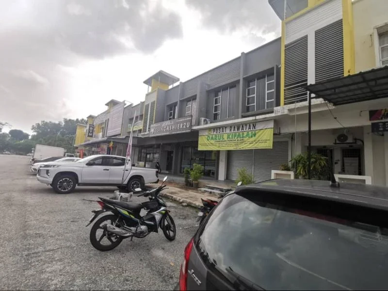 Exterior type: shophouse; features: grey painted facade, yellow accent panels, large glass windows, metal grilles, signage on facade, parking area, motorcycle parking, multiple storeys, surrounding gravel lot; style: modern.