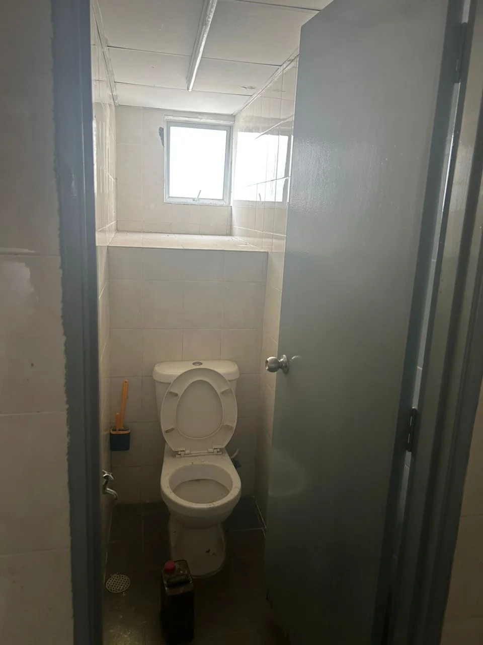 Bathroom features: white ceramic toilet, small window, white tiled walls, grey tiled flooring, metal door, toilet paper holder, cleaning supplies holder, sink with faucet, mirror above sink.