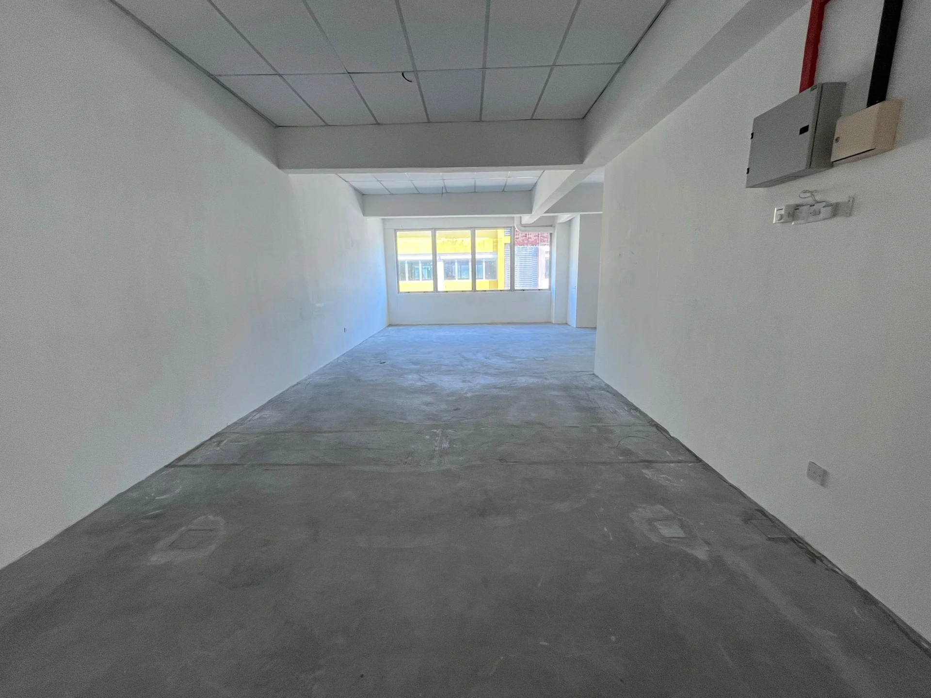 Room features: white painted walls, concrete flooring, ceiling with grid tiles, large window providing natural light, exposed electrical wiring, open layout, no visible fixtures or furniture; style: minimalist.