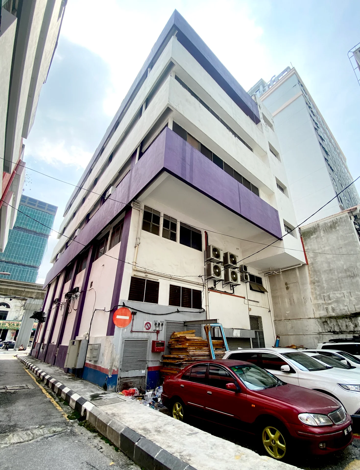 Exterior type: low-rise apartment block; features: purple painted facade, white painted facade, exposed air conditioning units, multiple storeys, parking area visible, surrounded by urban buildings, side alley access, signage visible, windows with grilles; style: modern.