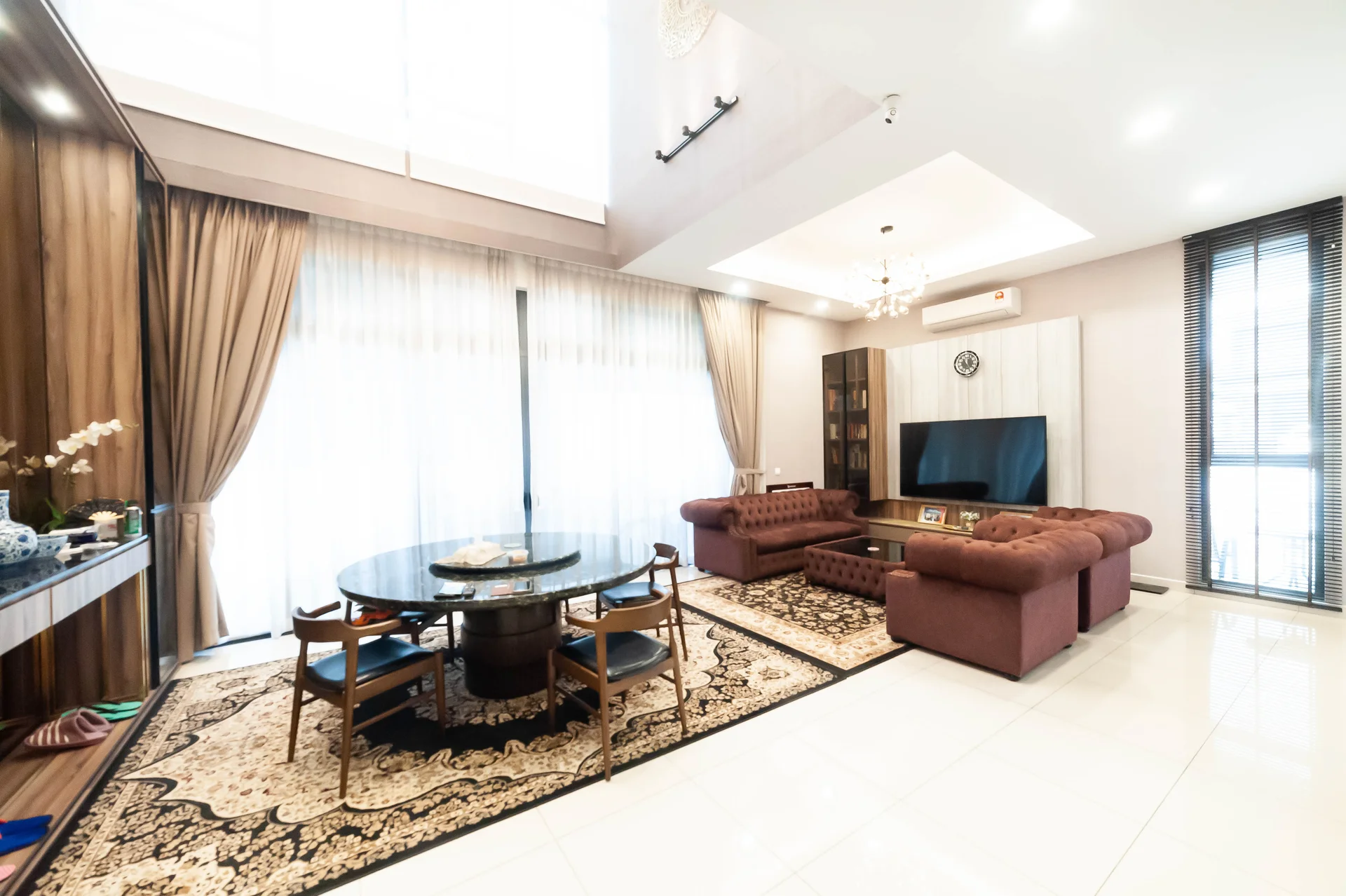 Living features: cream painted walls, white ceiling with recessed downlights, 5-blade ceiling fan with light, floor-to-ceiling windows with sheer curtains, dark wood cabinetry, large wall-mounted TV, brown leather sofa, round dining table with black marble top, upholstered dining chairs, area rug with intricate pattern, decorative wall clock, decorative plants in ceramic pots; style: modern.
