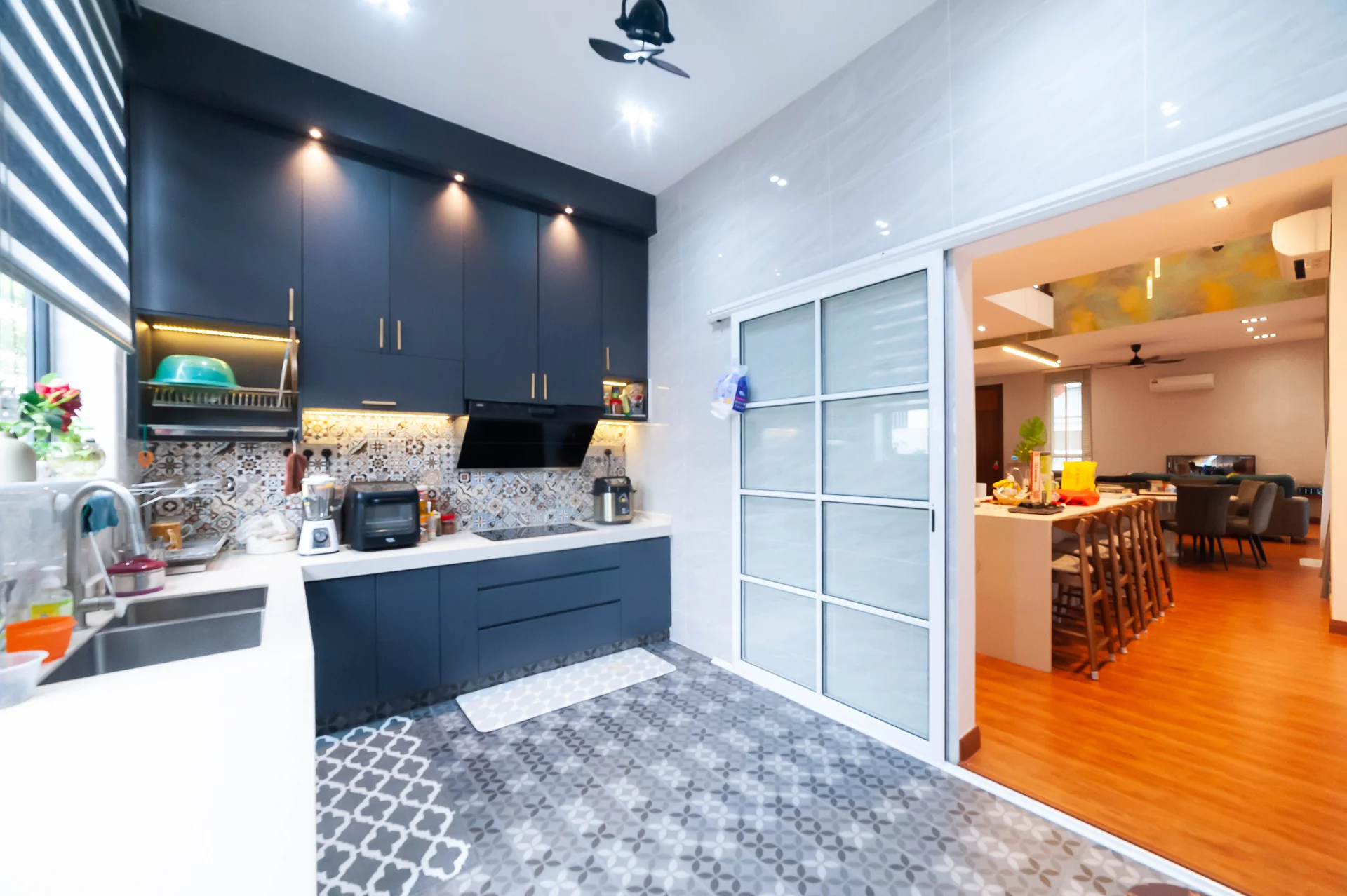 Kitchen features: dark blue cabinetry, white quartz countertop, single-bowl stainless-steel sink, chrome pull-down faucet, built-in oven, freestanding range oven, decorative backsplash with geometric pattern, recessed downlights, tile flooring with geometric pattern, window with sheer curtains, kitchen island, open layout to dining area; style: modern. Dining features: wooden dining table, high-backed dining chairs, open layout to kitchen, pendant lighting, wooden flooring; style: modern.