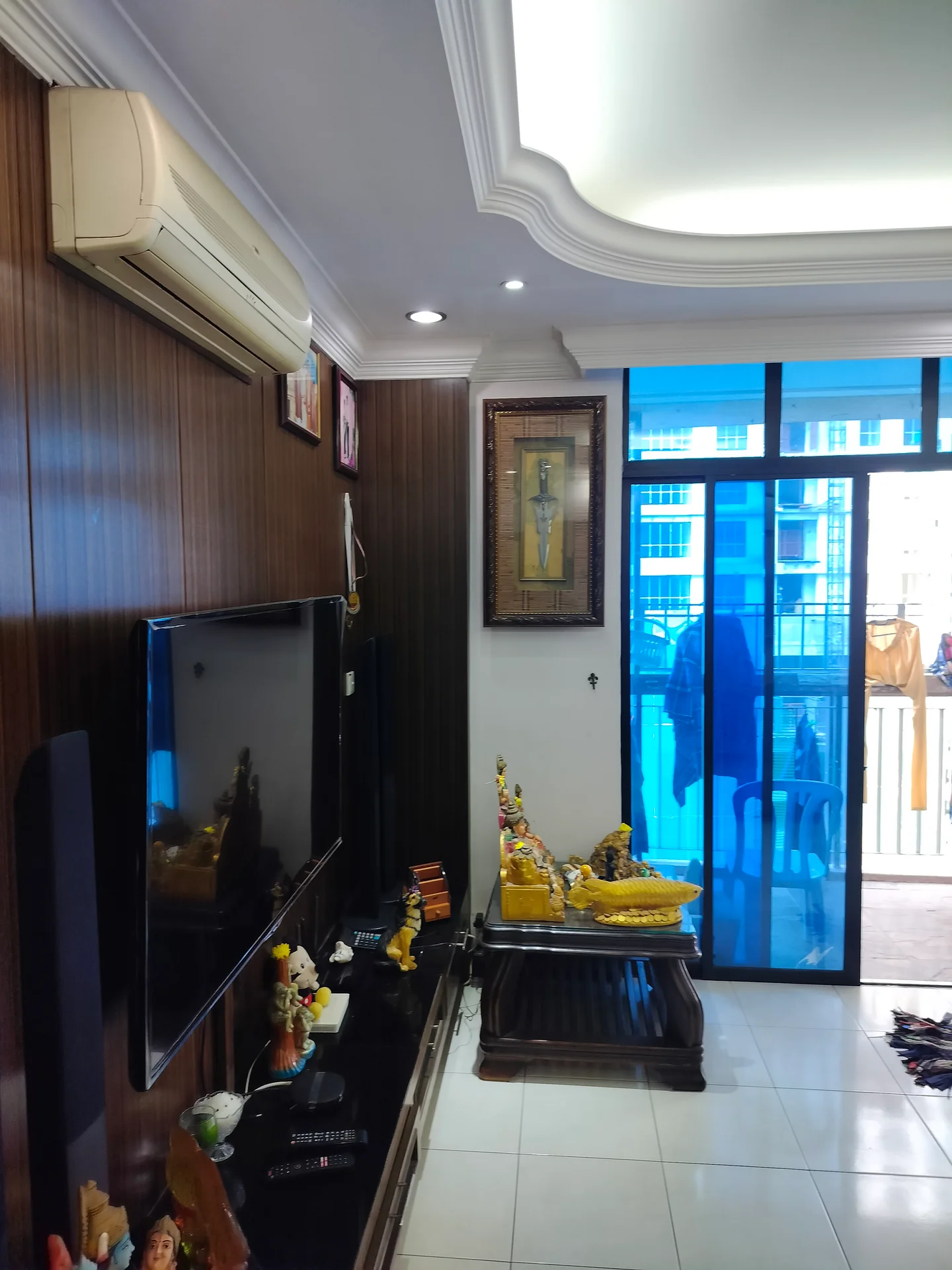 Living features: wall-mounted flat-screen TV, dark wood panelled wall, white ceiling with crown moulding, recessed downlights, 1 wall-mounted split AC, glass sliding door, white tiled flooring, decorative statue, wooden coffee table, framed artwork, decorative figurines; style: modern.