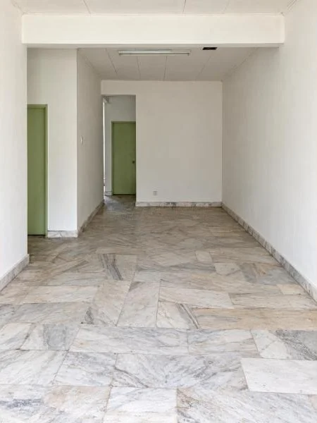 Hallway features: white painted walls, marble tile flooring, white ceiling, recessed fluorescent ceiling light, narrow rectangular layout, plain flat walls, green painted door frames, multiple interior door openings, baseboard trim, unfinished or vacant interior, natural light from unseen source; style: minimalist.