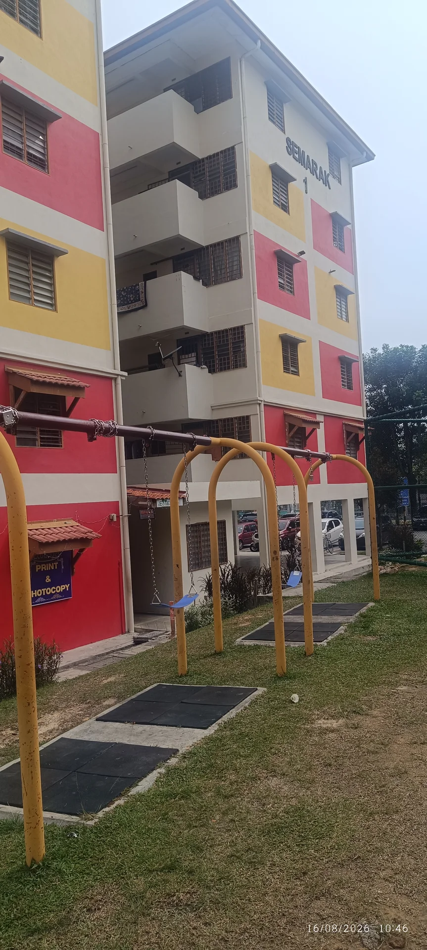 Exterior type: low-rise apartment block; features: red and yellow painted facade, 3-storey building, swing set in foreground, manicured lawn, sidewalk, parking area visible, windows with metal grilles; style: modern.