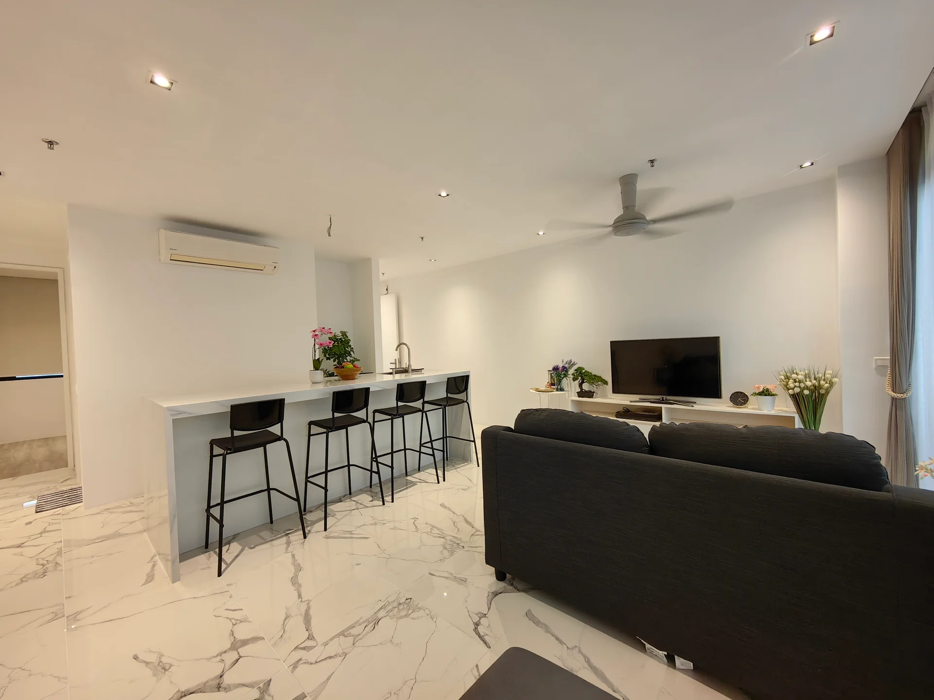 Living features: 3-seater fabric sofa in black, coffee table, wall-mounted TV, TV console, marble tile flooring, recessed downlights, 5-blade ceiling fan, floor-to-ceiling windows, decorative plants in ceramic pots, open shelving, bar counter with 3 black stools; style: modern.