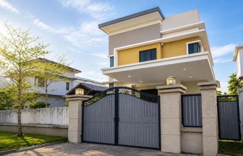 Exterior type: detached bungalow; features: double-storey, cream and grey painted facade, flat roof, projecting balconies, covered car porch, double-leaf metal gate, perimeter wall, gated entry, stone-clad gate pillars, driveway with concrete paving, manicured lawn, young tree, surrounded by residential houses; style: modern.