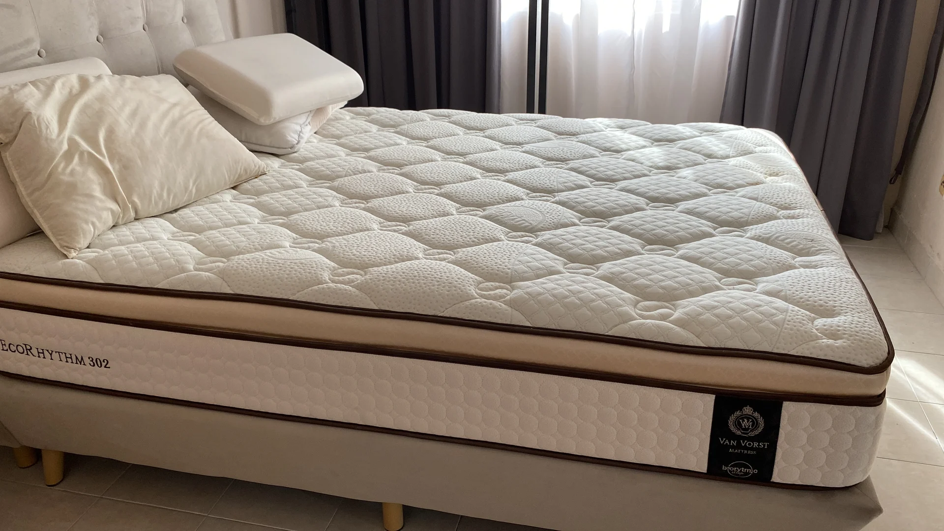 Bedroom features: queen-size mattress, white quilted mattress cover, decorative pillow, bed frame with light-colored upholstery, floor-to-ceiling window with sheer curtains, grey curtains, tile flooring, neutral wall color; style: modern.