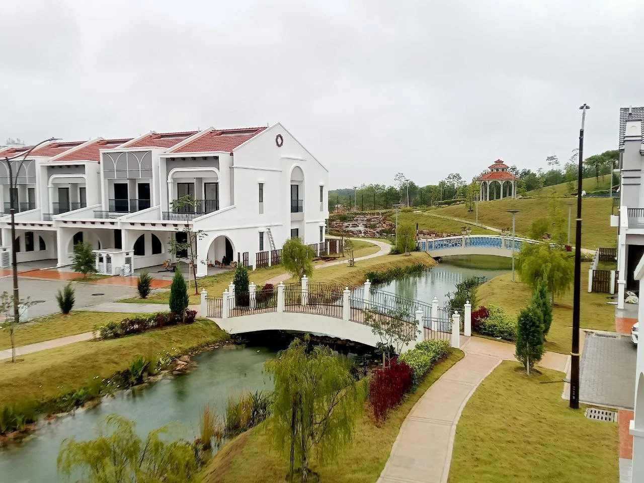 Exterior type: linked terrace house; features: white painted facade, red pitched tile roof, manicured lawn, landscaped garden, small bridge over water, water feature with pond, gazebo structure, pathway with interlocking pavers, surrounded by greenery; style: modern.