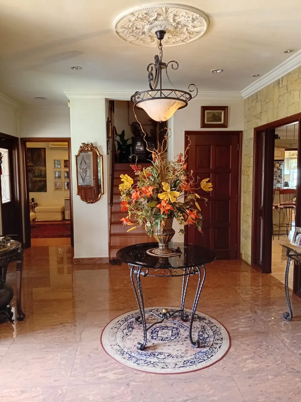 Hallway features: marble flooring, cream painted walls, exposed stone accent wall, chandelier with glass shade, decorative ceiling rose, round decorative table, floral arrangement in vase, framed artwork on wall, wooden door, staircase leading to upper floor; style: traditional.