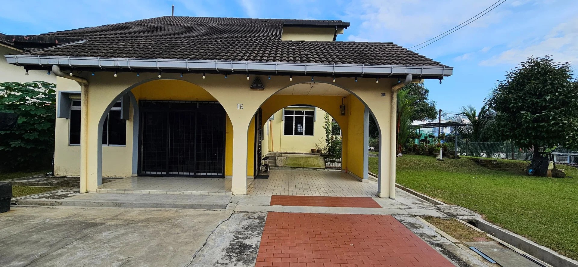 Exterior type: detached bungalow; features: yellow painted facade, pitched tile roof, arched entrance with columns, paved walkway, manicured lawn, garden area, visible windows, security grille at entrance; style: modern.