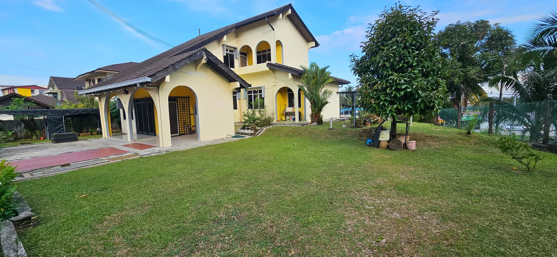 Exterior type: detached bungalow; features: yellow painted facade, pitched tile roof, arched windows, manicured lawn, mature trees, paved walkway, garden area, outdoor seating area, fenced perimeter, surrounded by greenery; style: traditional.