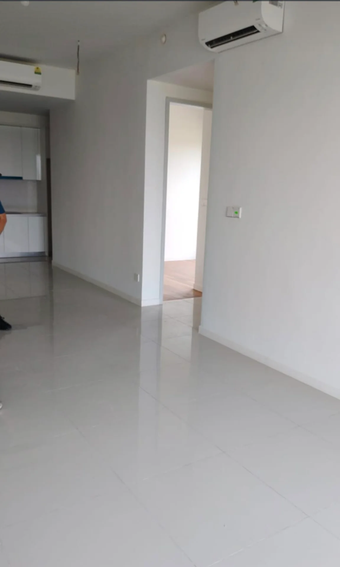 Room features: white painted walls, glossy ceramic tile flooring, 1 wall-mounted split AC, recessed ceiling lights, doorway leading to another room, kitchen area visible in background, neutral color scheme; style: modern.