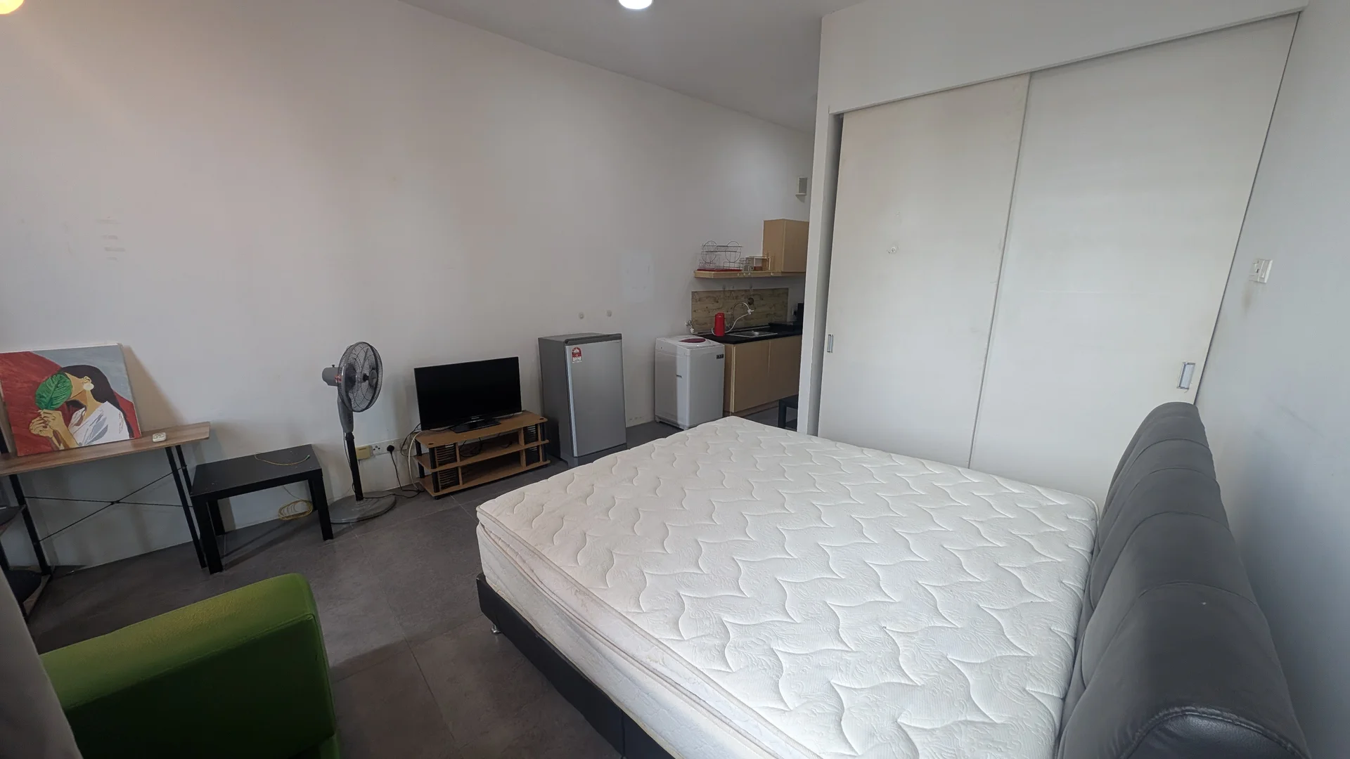 Bedroom features: queen-size bed, mattress without bedding, grey upholstered headboard, small study desk, black chair, fan, fridge, kitchenette with countertop, built-in wardrobe, grey tiled flooring, white painted walls, ceiling light; style: modern.