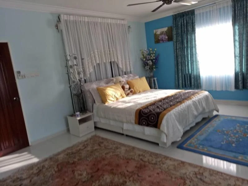 Bedroom features: king-size bed with white bedding, upholstered headboard, two yellow decorative pillows, white bedside table, area rug with floral pattern, blue area rug, ceiling fan, curtains with floral pattern, wall-mounted artwork, decorative flowers in vase, light blue painted walls, white ceiling, natural light from window; style: contemporary.
