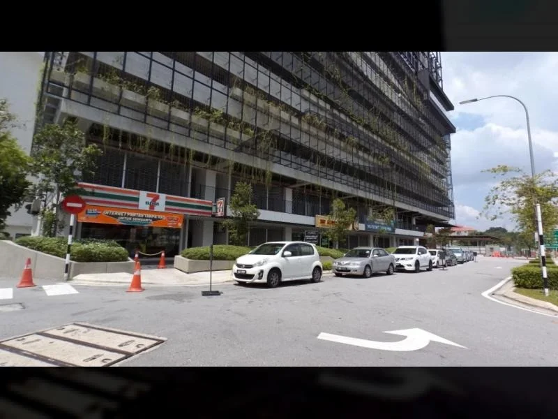 Exterior type: high-rise condominium; features: glass curtain wall, greenery on balconies, multiple retail units at ground level, surrounded by landscaped areas, driveway with interlocking pavers, parking spaces visible, signage for convenience store, traffic cones for parking management; style: modern.