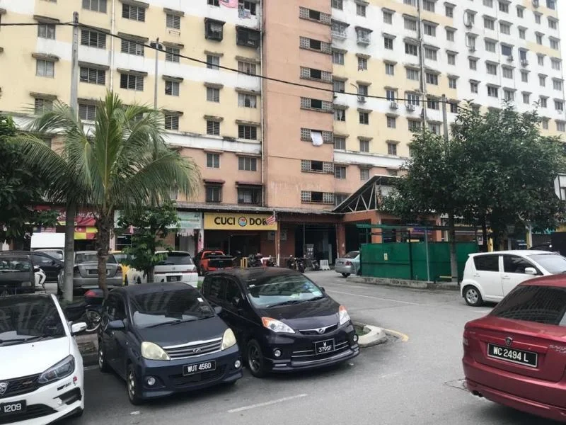 Exterior type: low-rise apartment block; features: multi-storey building, concrete facade, balconies visible, ground-level parking area, palm trees in foreground, surrounded by greenery, entrance with awning, visible signage for laundry service; style: modern.