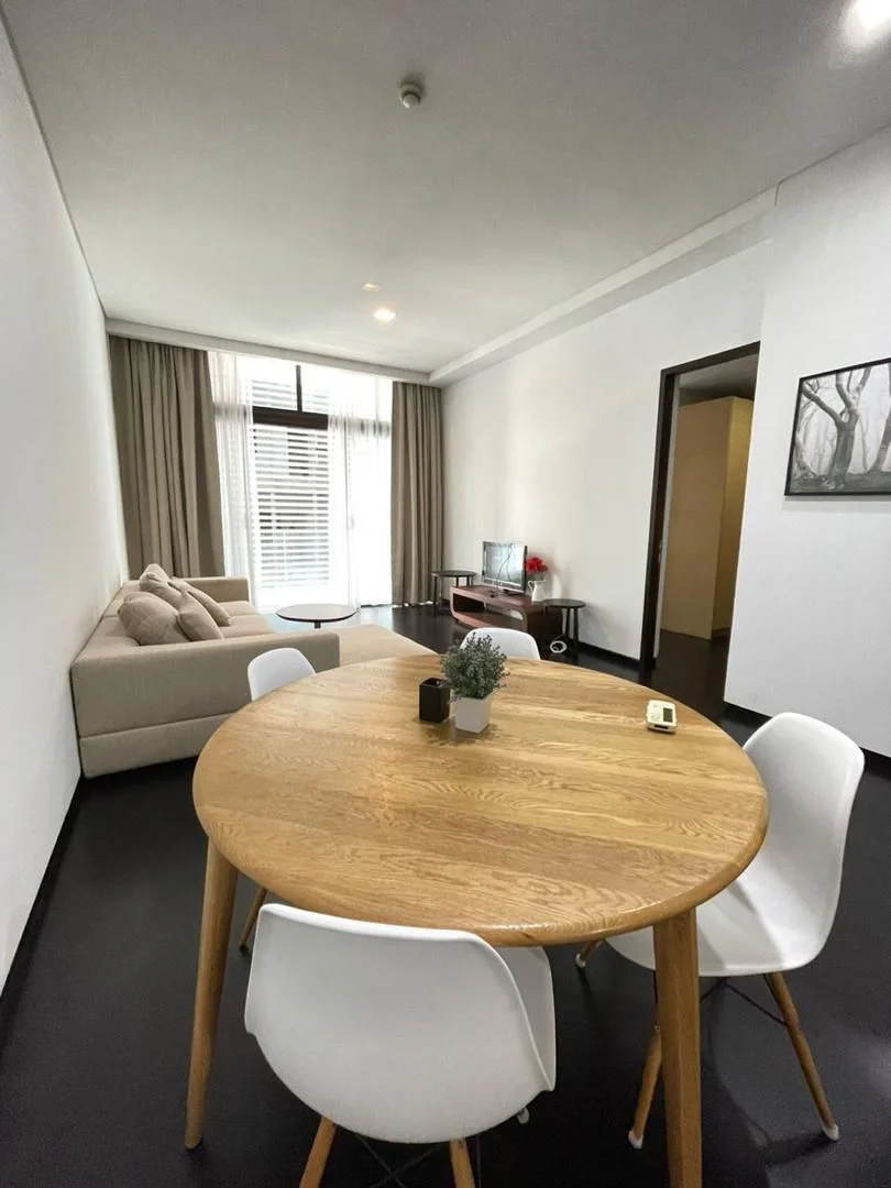 Living features: light grey fabric sofa, round wooden dining table, white plastic dining chairs, floor-to-ceiling windows, sheer curtains, dark wood flooring, TV console, wall-mounted TV, decorative plant on table, framed artwork on wall; style: modern.