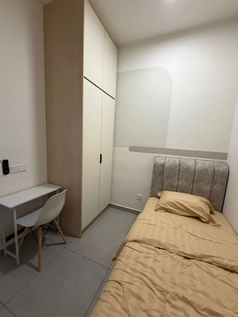 Bedroom features: single bed with beige bedding, upholstered headboard in grey, built-in wardrobe with sliding doors, white desk with wooden legs, white chair, grey tiled flooring, white painted walls, wall-mounted light switch, power outlets; style: modern.