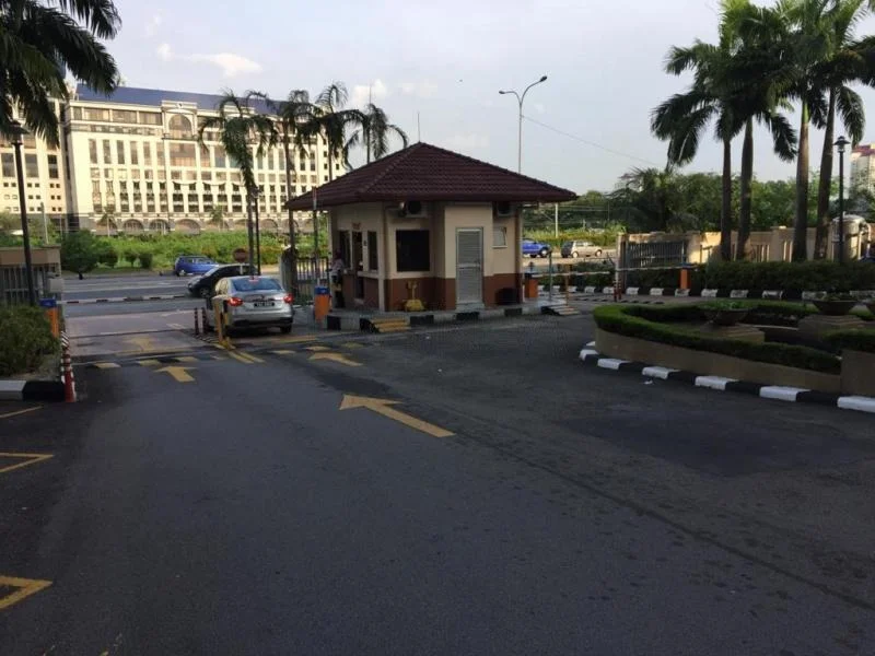 Exterior type: main entrance gate; features: guard house at entrance, paved driveway, palm trees, landscaped greenery, security boom gate, marked parking spaces, surrounding road, adjacent building in background; style: modern.