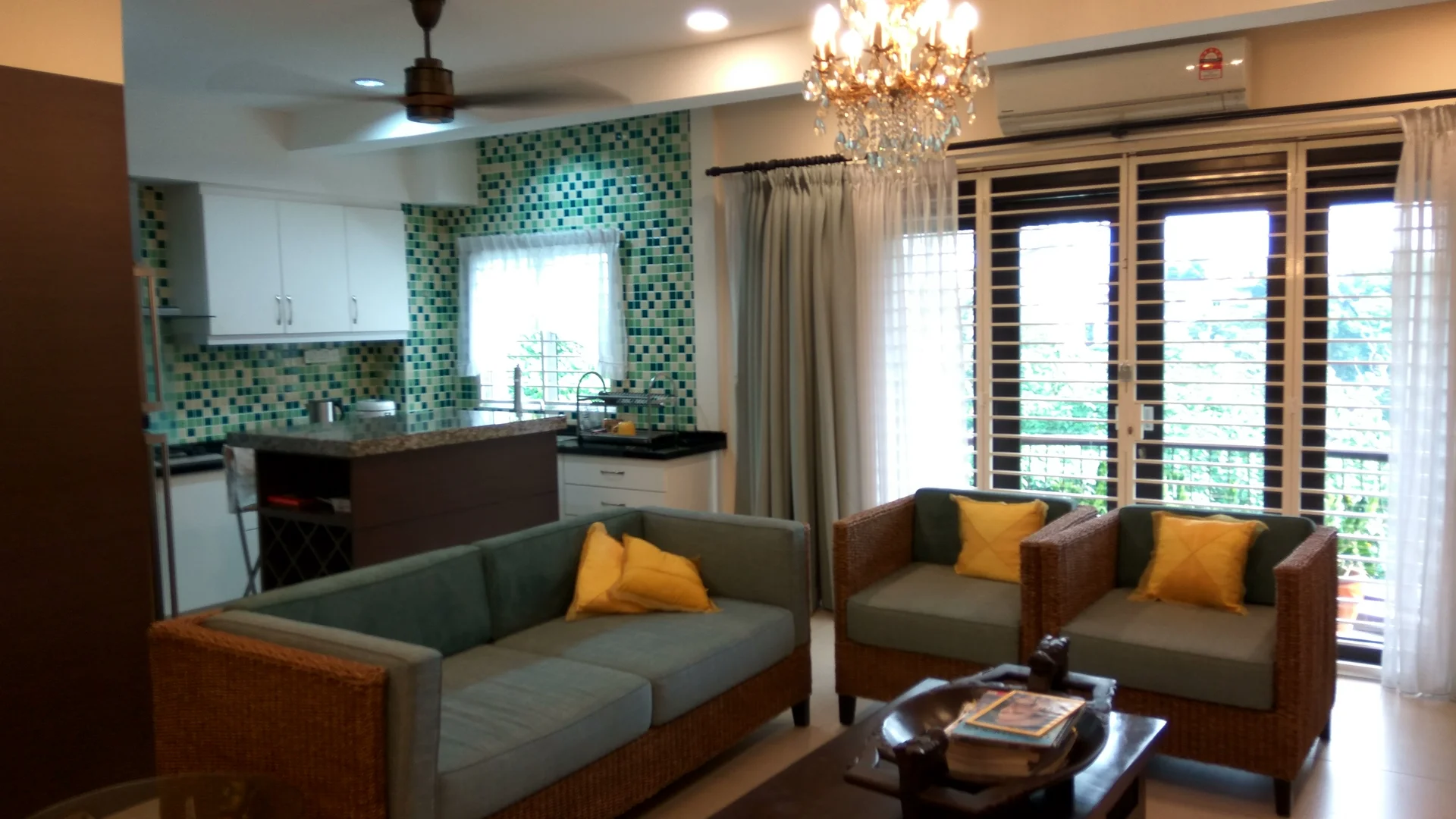 Living features: 3-seater fabric sofa in light blue, 2-seater rattan sofa with cushions, yellow decorative cushions, wooden coffee table, chandelier with crystals, ceiling fan, floor-to-ceiling windows with horizontal blinds, green mosaic tile backsplash, white cabinetry, black countertop, kitchen island with bar seating, open layout to kitchen, natural light from windows, decorative wall art; style: modern; view: garden.