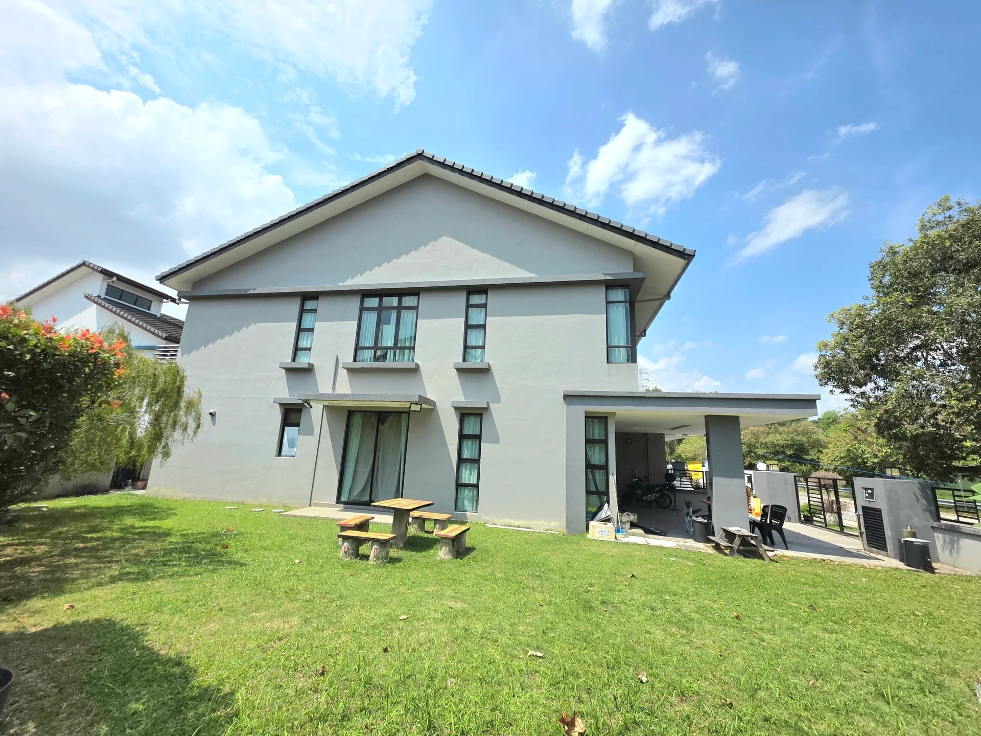 Exterior type: detached bungalow; features: grey painted facade, flat roof, two-storey building, large windows, covered patio area, manicured lawn, outdoor seating area with picnic table, surrounded by greenery, driveway, fenced perimeter; style: modern.