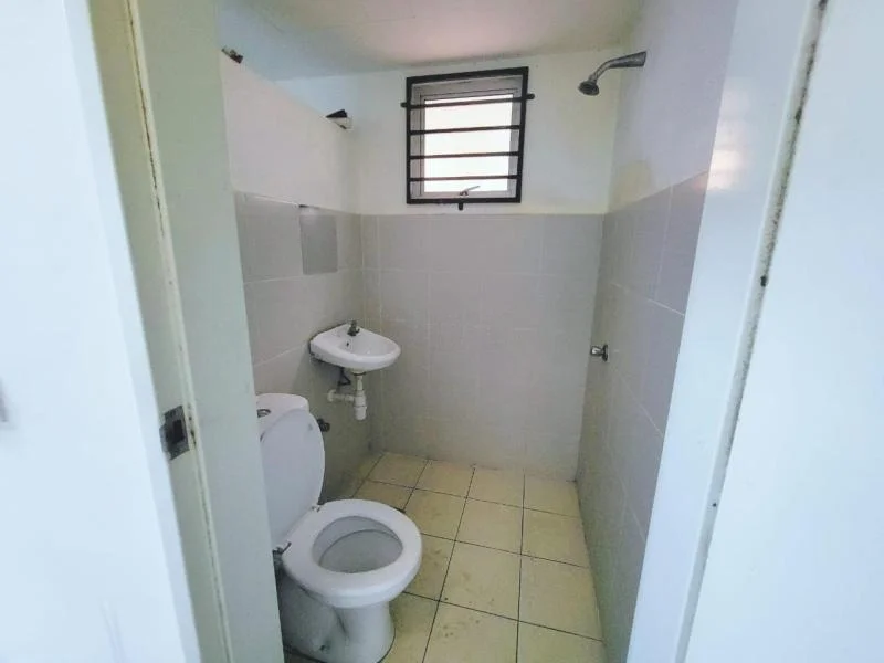 Bathroom features: white tiled walls, light grey tiled flooring, one-piece toilet, wall-mounted sink, shower head, window with metal grille, chrome faucet.
