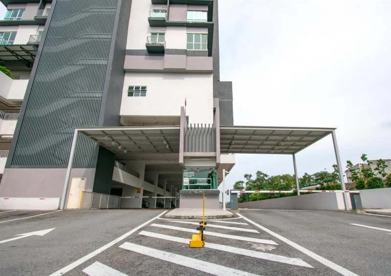 Exterior type: mid-rise condominium; features: covered parking area, driveway with marked lanes, security barrier at entrance, multiple levels of parking, modern architectural design, grey and white facade, balconies visible on upper floors, landscaped surroundings; style: modern.