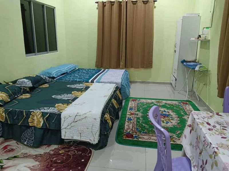 Bedroom features: two beds with floral bedding, green walls, white tiled flooring, curtains in brown, window with glass panes, small white cabinet, table with floral tablecloth, purple plastic chairs, area rug with floral pattern.