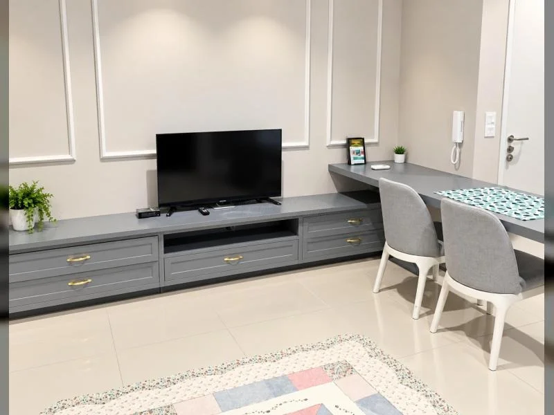 Living features: white painted walls, decorative wall moulding, glossy beige tile flooring, recessed ceiling downlights, low-profile grey TV console with drawers, flat-screen TV, set-top box, remote controls, small potted plant, built-in desk nook, grey desktop surface, two grey upholstered dining chairs, wall-mounted intercom handset, white interior door, gold-tone drawer handles, area rug with pastel geometric pattern; style: contemporary.