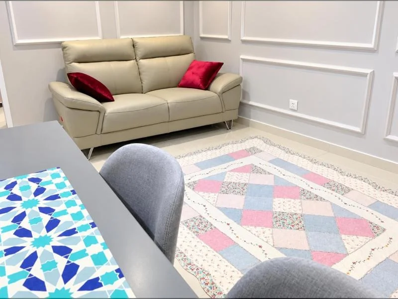 Living features: beige 2-seater leather sofa, red throw pillows, light grey painted walls, decorative wall moulding panels, cream glossy tile flooring, pastel patchwork area rug, grey upholstered dining chair, grey rectangular table, patterned blue and turquoise table runner, white electrical outlet, minimalist contemporary decor; style: contemporary.