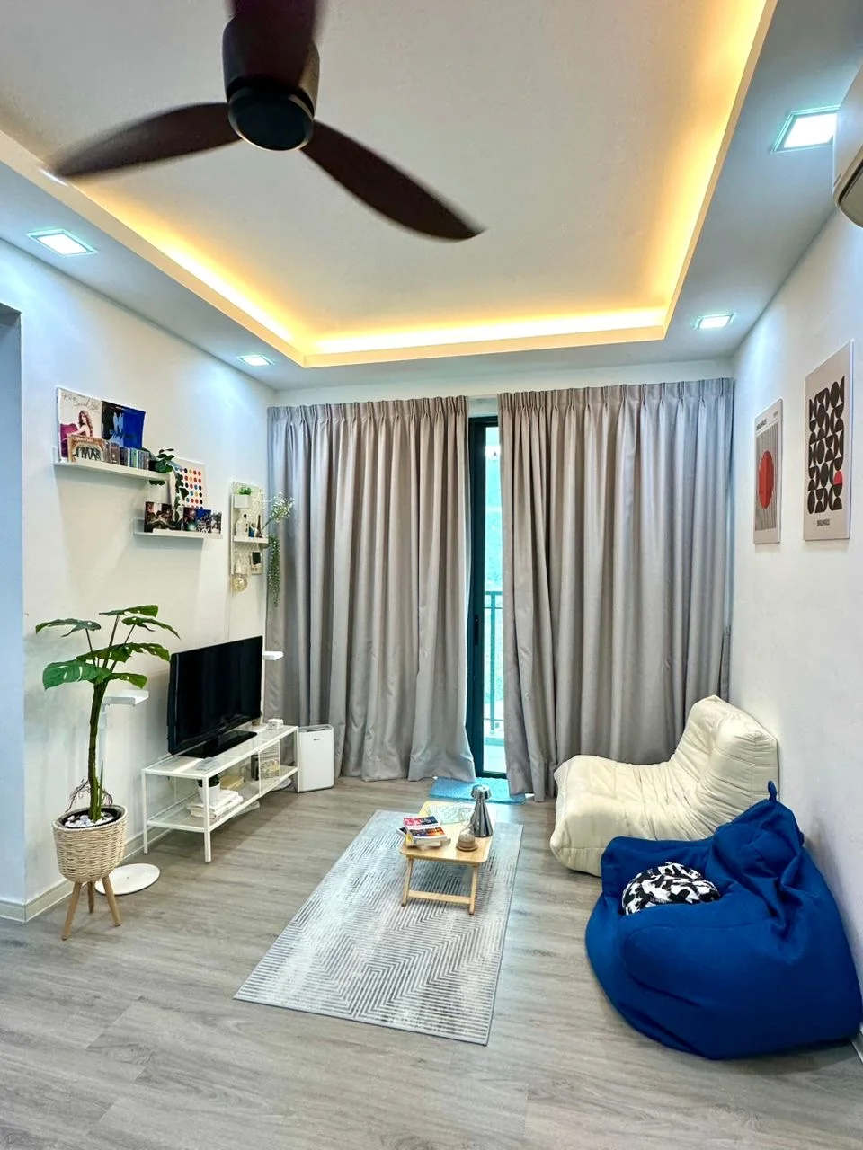 Living features: 5-blade ceiling fan with light, recessed LED downlights, floor-to-ceiling windows, grey curtains, white fabric sofa, blue bean bag chair, TV console, wall-mounted TV, decorative wall art, area rug with geometric pattern, potted plant, coffee table; style: modern.