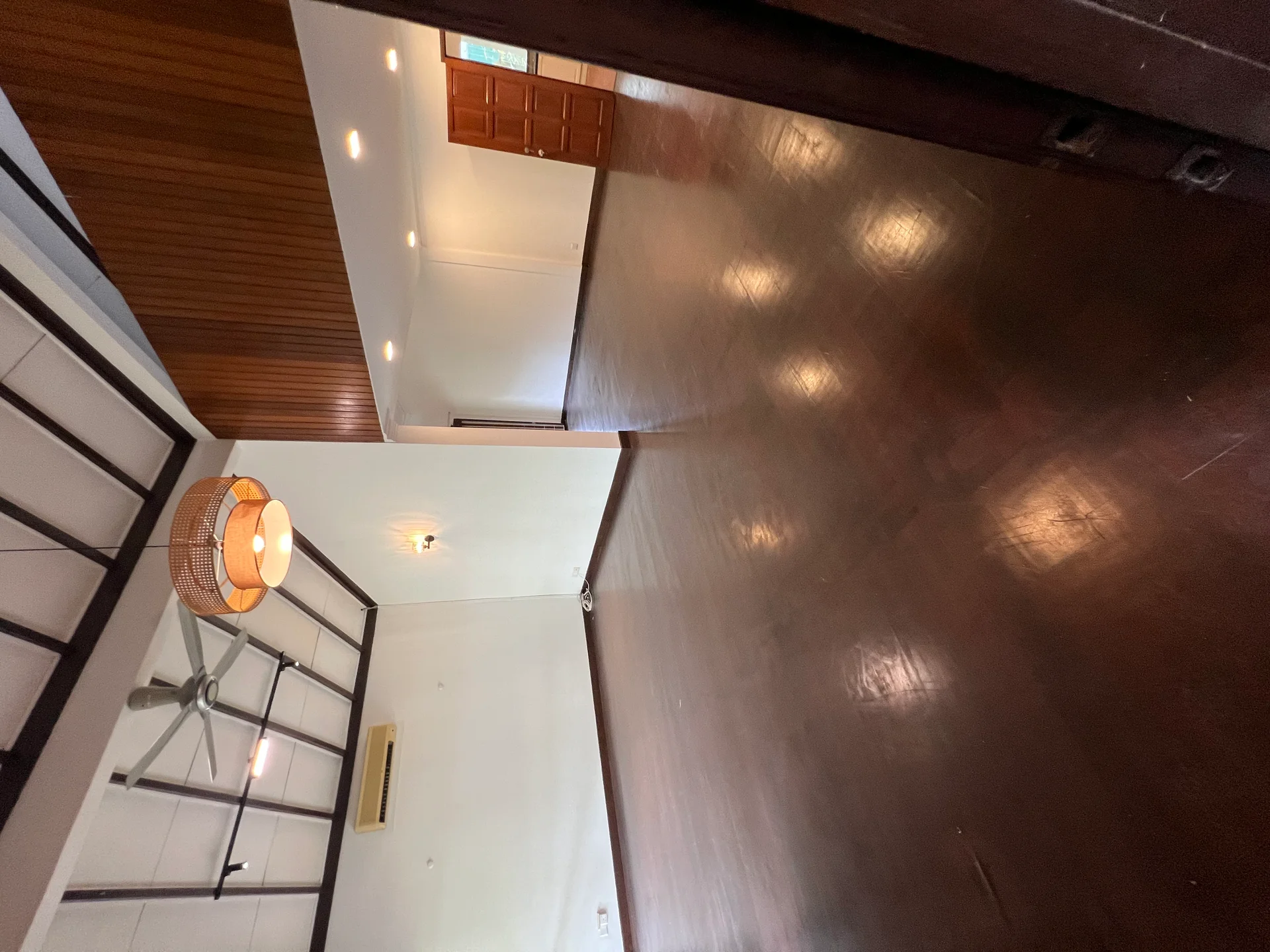 Living features: wooden flooring, exposed beams on ceiling, 5-blade ceiling fan, pendant light, white painted walls, large open space, natural light from windows, air conditioning unit; style: modern.