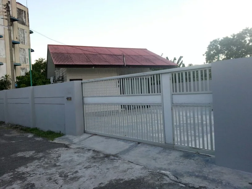 Exterior type: detached bungalow; features: grey painted facade, red pitched tile roof, metal gate, concrete driveway, surrounded by low wall, manicured lawn, mature trees, paved walkway; style: traditional.