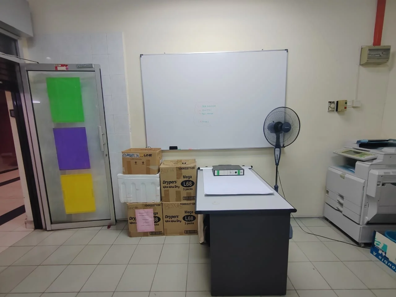 Room features: whiteboard, ceiling fan, printer, office desk, cardboard boxes, refrigerator with colored panels, tiled flooring, wall-mounted electrical outlet.