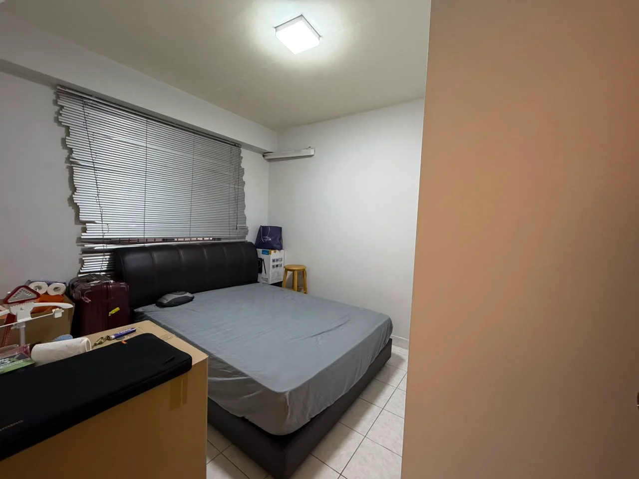 Bedroom features: queen-size bed with grey bedding, black upholstered headboard, white walls, tile flooring, window with horizontal blinds, wall-mounted split AC unit, small stool, luggage, nightstand, light fixture on ceiling; style: modern.