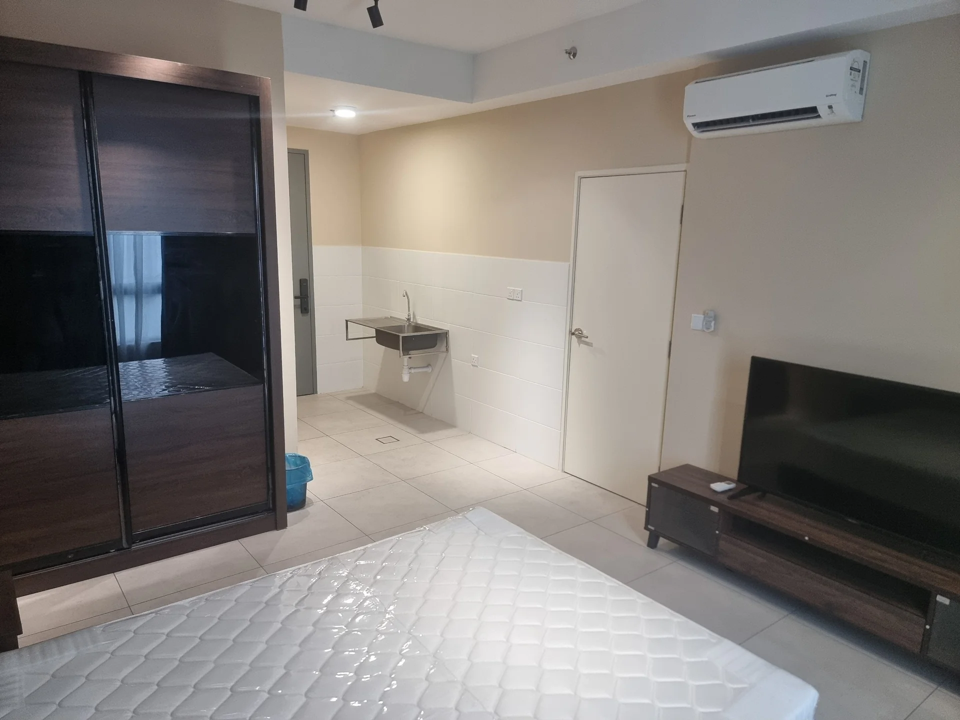 Room features: wall-mounted split AC, tile flooring, light beige painted walls, built-in wardrobe with sliding doors, TV console, wall-mounted TV, sink with faucet, white tiled backsplash, door to bathroom, recessed lighting; style: modern.