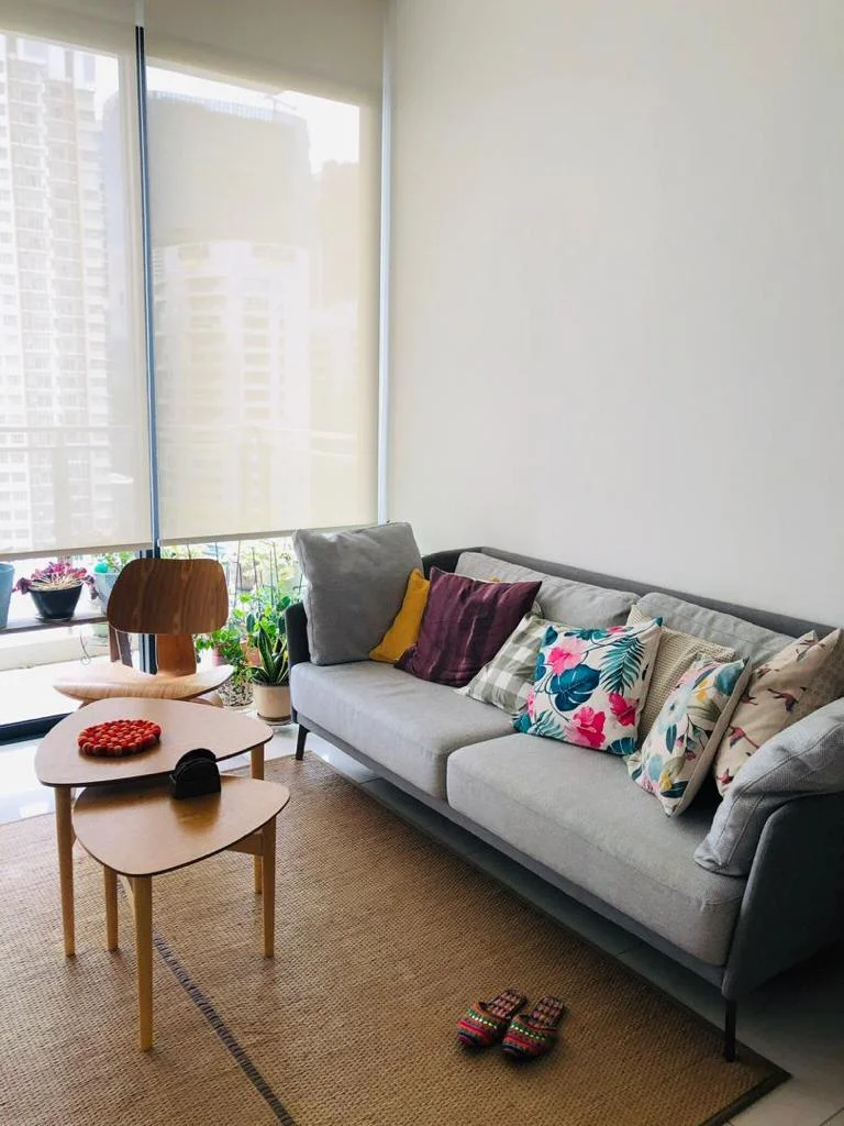 Living features: grey fabric 3-seater sofa, decorative cushions with floral and geometric patterns, wooden coffee table, woven area rug, floor-to-ceiling windows, roller blinds, indoor plants, slippers on the floor; style: modern; view: city.